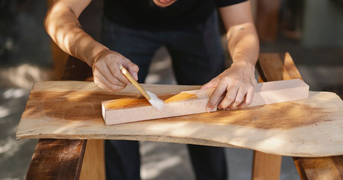 A brush laying a coat of finish along a planed length of pale hardwood resting on a live-edge slab.